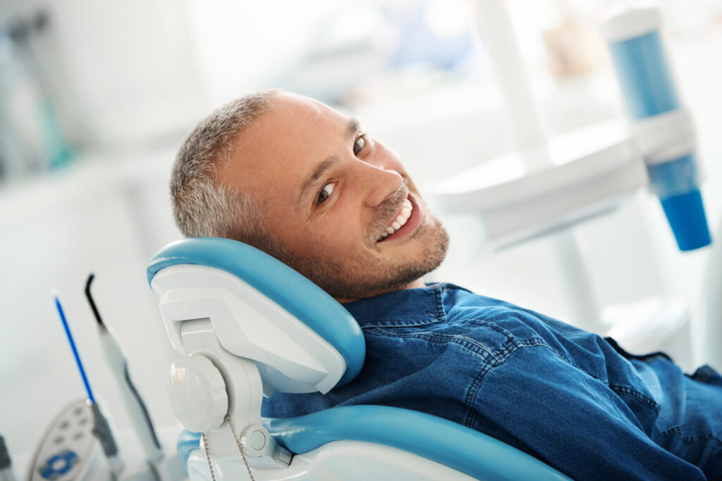 Mature man with short hair in a jean jacket smiling while laying back in the dental treatment chair